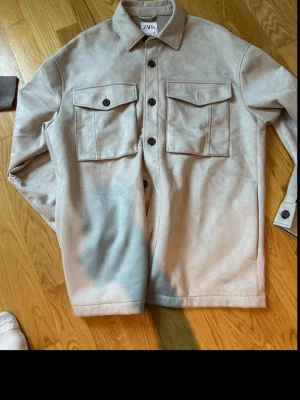 Beige overshirt från Zara - Stilren beige overshirt från Zara med två stora bröstfickor och knappar framtill. Skjortjackan har klassisk krage och är tillverkad i ett mjukt material som ger en avslappnad look. Perfekt att bära över en t-shirt för en trendig lager-på-lager stil.
