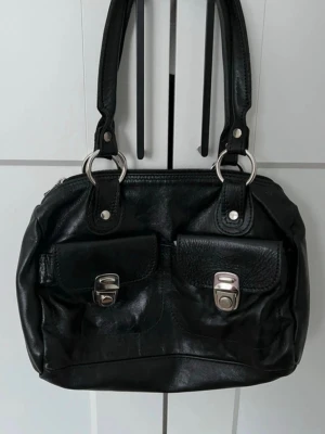 Svart handväska med dubbla fickor - Snygg svart handväska i skinnimitation med två framfickor som har silverfärgade spännen. Väskan har dubbla handtag och stora silverringar som detaljer. Stängs med dragkedja upptill och har en extra ficka på baksidan. Perfekt för dig som gillar en stilren och praktisk look.