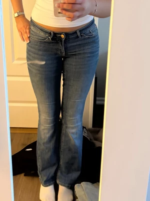 Blå bootcut jeans med låg midja - Säljer ett par klassiska blå bootcut jeans med låg midja och fem fickor. Jeansen har en gul knappdetalj och subtila slitningar framtill för en avslappnad look. Perfekta till sneakers eller boots och passar dig som gillar en tidlös stil.