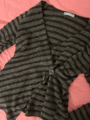 Randig kofta från LoralKitsch - Mysig kofta i svart och grått med horisontella ränder från LoralKitsch. Koftan har en knapp framtill, asymmetrisk nederkant och långa ärmar. Materialet är mjukt och har en lätt glittrig finish som ger extra stilpoäng.