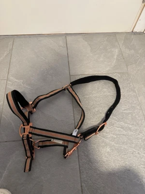 Svart och beige grimma till häst - Säljer en grimma i svart och beige med detaljer i roséguld. Grimman har justerbara spännen och är tillverkad i slitstarkt nylonmaterial. Passar perfekt till häst och har mjuk vaddering vid nacken för extra komfort.