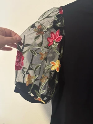 2XL, oanvänd Svart topp med broderade blommiga ärmar - Svart topp från Shein med transparenta puffärmar i mesh och färgglada broderade blommor i rött, gult och grönt. Kroppen är enkel och slät medan ärmarna ger en unik och trendig look. Perfekt för dig som vill sticka ut lite extra.
