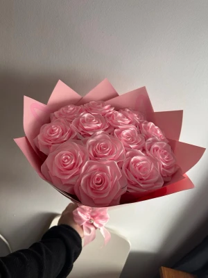 Bukett med rosa konstgjorda rosor - Säljer en bukett med konstgjorda rosor i ljusrosa färg. Rosorna är gjorda av ett glansigt material och är arrangerade i ett dekorativt rosa omslagspapper med rosett. Perfekt som present eller dekoration.