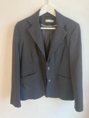 Svart kavaj från WERA - Snygg mörkgrå kavaj från Vera med tunna vita kritstrecksränder. Klassisk modell med två knappar framtill, spetsiga slag och tre fickor. Kavajen har helfodrad insida och fyra dekorativa knappar vid ärmsluten. Perfekt för en stilren look.