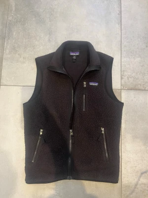 Svart fleeceväst från Patagonia  - Mysig svart fleeceväst från Patagonia med hög krage och tre dragkedjeförsedda fickor framtill. Västen har en klassisk rak passform och är tillverkad i mjukt teddyfleece-material. Perfekt att slänga över en hoodie eller tröja för extra värme.