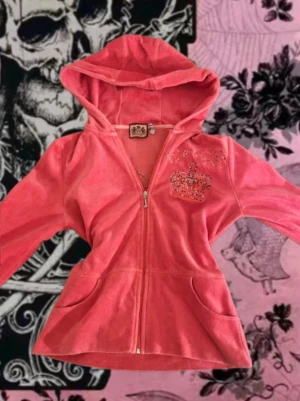 Rosa Juicy Couture hoodie - Juicy Couture hoodie i mjuk rosa material med dragkedja och huva. Framsidan har en diamant detaljerad krona och på ryggen finns en större diamant detaljerad krona med text där det står juicy couture. Två fickor framtill och figurnära passform. Perfekt för en avslappnad men stylish look.