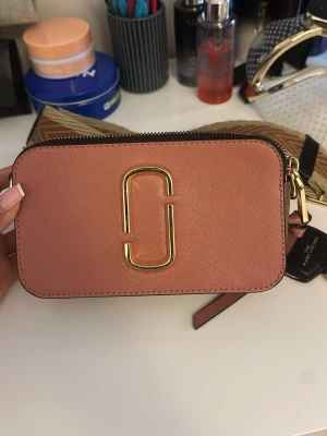 Rosa Marc Jacobs Snapshot handväska - Säljer en snygg rosa Marc Jacobs Snapshot handväska i strukturerat läder med guldfärgad logga framtill. Väskan har en rektangulär form, dragkedja upptill och avtagbar axelrem. Perfekt för dig som gillar trendiga accessoarer och vill ha något som sticker ut.
