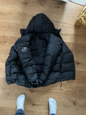 Svart dunjacka från sail racing - Snygg svart dunjacka från Sail racing med huva och dragkedja framtill. Jackan har quiltad design, logotyp på ärmen och praktiska fickor. Perfekt för kalla vinterdagar och riktigt varm tack vare dunfyllningen.