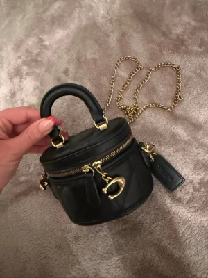 Coach svart minihandväska i läder - Supersöt svart minihandväska från Coach i quiltat läder med guldfärgade detaljer och kedjerem. Väskan har rund form, dragkedja upptill och ett dekorativt C-spänne framtill. Perfekt för det viktigaste och ger en lyxig touch till din outfit.