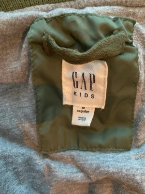 Olivgrön bomberjacka från GAP Kids - Jätte Snygg olivgrön bomberjacka från GAP Kids med dragkedja framtill, två fickor med knapp och en ärmficka med dragkedja. Jackan har ribbade muddar vid ärmslut och midja. Tillverkad i slitstarkt material, perfekt för en cool och avslappnad stil. Motsvarande storlek 130. Namnad men lapp går att klippa bort
