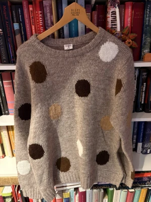 Grå stickad tröja med prickar Stories - Mysig grå stickad tröja från Stories i storlek M, dekorerad med stora prickar i brunt, vitt, beige och svart. Tröjan har rund halsringning och ribbade muddar. Perfekt för dig som gillar lekfulla detaljer och en avslappnad stil.