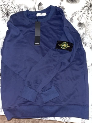 Blå sweatshirt från Stone Island - Snygg blå sweatshirt från Stone Island med klassisk rund hals och ribbade muddar. Tröjan har den ikoniska Stone Island-loggan på vänster ärm och är tillverkad i mjukt bomullsmaterial. Perfekt för en avslappnad och trendig stil.