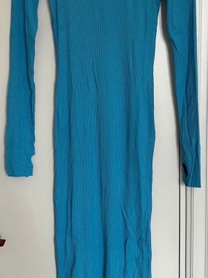 Turkos ribbad långärmad klänning - Säljer en turkos, ribbad långärmad klänning i stretchigt material. Klänningen har en kroppsnära passform och hög hals, perfekt för dig som gillar en enkel men snygg stil. Materialet känns mjukt och följsamt mot kroppen.