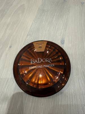 IsaDora Bronzing Powder Golden Tan 46 - IsaDora Bronzing Powder i nyansen Golden Tan 46. Kommer i en rund, brun plastförpackning med spegel i locket. Ger en solkysst look med sin varma, gyllene ton. Perfekt för att framhäva ansiktets konturer och ge glow.