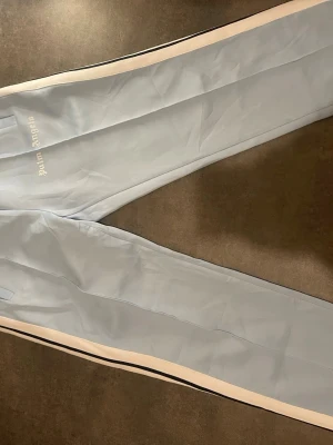 Ljusblåa mjukisbyxor Palm Angels - Snygga ljusblåa mjukisbyxor från Palm Angels med vita sidoränder och logga broderad på låret. Byxorna har resår i midjan och är tillverkade i ett mjukt, sportigt material. Perfekta för en chill och trendig look.