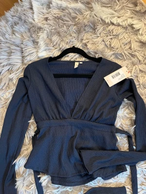 Mörkblå omlottopp från Nelly - Trendig mörkblå omlottopp från Nelly med lång ärm och v-ringning. Toppen har ribbad struktur och ett knytband i midjan som ger en snygg siluett. Perfekt att styla med jeans eller kjol för en cool look.