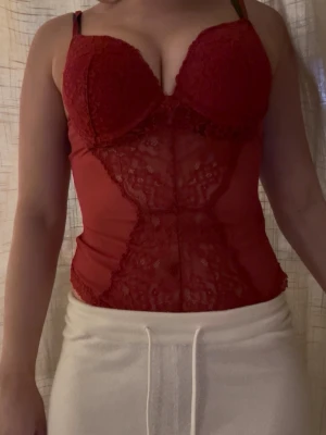 Röd spetsbody med meshdetaljer - Snygg röd body med vadderade kupor och tunna axelband. Framsidan har genomskinliga meshpartier och dekorativa spetsdetaljer som ger en feminin look. Perfekt för dig som vill ha något extra under kläderna. Materialet är stretchigt och följsamt.