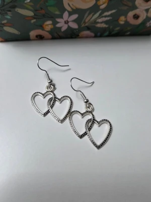 Silvriga örhängen med dubbla hjärtan i Rostfritt Stål Nya - Söta örhängen med två sammanflätade hjärtan på varje krok. ✨ Handgjorda av mig ✔️ Material: Rostfritt stål – tåliga och allergivänliga ✔️ Färg: Silver ✔️ Skick: Nya / oanvända Perfekta som present eller till vardag/fest 💖 Frakt: Samfraktar gärna.