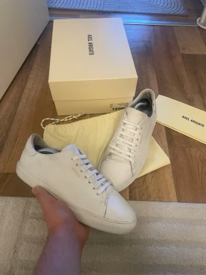 Vita Axel Arigato sneakers i läder - Stilrena vita sneakers från Axel Arigato i mjukt läder. Klassisk låg modell med vita skosnören och diskret logga på sidan. Perfekta för dig som gillar minimalistisk och clean stil. Kommer med originalkartong och dustbag.obs box medföljer ej 