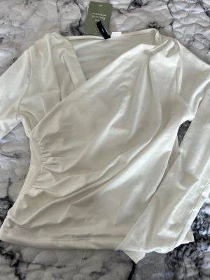 Vit omlottopp från H&M - Säljer en stilren vit omlottopp från H&M med lång ärm och lätt rynkad detalj i sidan. Toppen har en v-ringad omlottdesign och är gjord i mjuk bomullsmix som känns skön mot huden. Perfekt att matcha med jeans eller kjol för en trendig look.