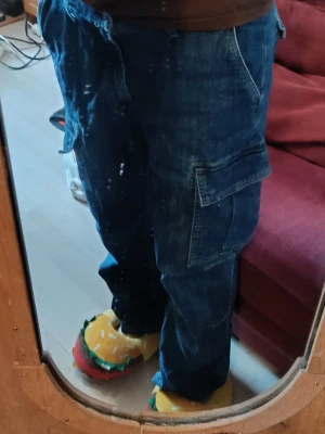 Blå cargobyxor med fickor - Säljer ett par blå cargobyxor i denim med flera stora fickor på sidorna. Byxorna har avslappnad passform och klassiska cargodetaljer som sömmar och ficklock. Perfekta för dig som gillar streetwear och vill ha både stil och funktion. Jag har själv sytt in byxorna i midjan för att dom skulle passa men om du tar bort stygnen blir dom en medium.