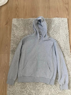 Grå zip hoodie med huva - Klassisk gråmelerad hoodie med dragkedja framtill och två fickor. Tröjan har en rymlig huva med snörhål och ribbade muddar vid ärmar och nederkant. Perfekt för en avslappnad och enkel stil. Materialet känns mjukt och bekvämt, troligen bomullsblandning.