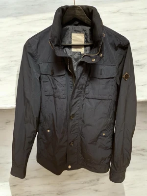 Mörkblå J.Lindeberg jacka - Stilren svart vindjacka från J.Lindeberg med hög krage, fyra framfickor med knappstängning och detaljer i metall. Jackan har en rak passform och är tillverkad i ett lätt, vindtätt material som passar perfekt för blåsiga dagar. St XS, sparsamt använd