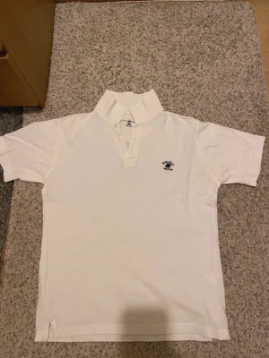 Vit pikétröja från Beverly Hills Polo Club - Klassisk vit pikétröja från Beverly Hills Polo Club med korta ärmar och knappar vid halsen. Tröjan har en broderad logga på bröstet och är tillverkad i mjuk bomull. Perfekt till en avslappnad och stilren look.