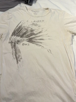 Vit t-shirt från Denim & Supply Ralph Lauren - Vit t-shirt från Denim & Supply Ralph Lauren med ett stort grafiskt tryck av en fjäderprydd huvudbonad på framsidan. Klassisk rund halsringning och korta ärmar. Tillverkad i mjuk bomull för en skön känsla. Bra skick. Pris går att diskutera