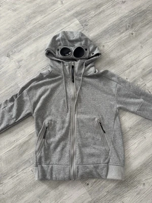 Grå hoodie med glasögon från C.P. Company - Säljer en grå hoodie från C.P. Company med ikoniska inbyggda glasögon i huvan. Tröjan har dragkedja framtill, två snedställda fickor med dragkedja och justerbar huva med snören. Tillverkad i mjuk bomullsblandning för maximal komfort. Storlek M