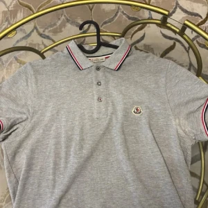 Grå pikétröja från Moncler - Snygg grå pikétröja från Moncler med klassisk krage och knäppning framtill. Tröjan har röda och vita detaljer på krage och ärmslut samt Moncler-logga broderad på bröstet. Perfekt för en stilren look.