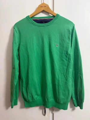 Grön stickad tröja från Gant - Snygg grön stickad tröja från Gant med rund halsringning och diskret broderad logga på bröstet. Tröjan har långa ärmar och ribbade muddar vid ärmslut och nederkant. Perfekt för dig som gillar klassisk och stilren design.