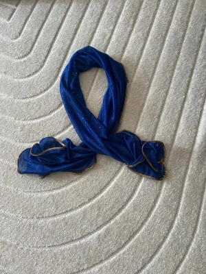 Blå glittrig sjal med guldkant - Snygg blå sjal i tunt, glittrigt tyg med skimrande guldkant runtom. Perfekt accessoar för att lyfta din outfit med lite extra färg och bling. Materialet känns lätt och luftigt, och sjalen har en rektangulär form som gör den enkel att styla på olika sätt.