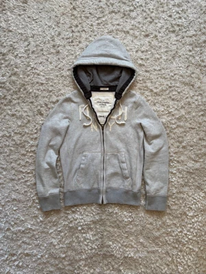 Abercrombie & fitch New York grå zip hoodie  - Sjukt snygg i trendig zip hoodie från Abercrombie & fitch. Den är använd och har defekter som syns på de sista bilderna, dessa gör det lite svårare att stänga hoodie än vanligt men är enligt mig inget stort problem. Storlek S, tveka inte på o skriva vid fler frågor! Längd: 65 Bröstvidd: 54 Ärm: 68 