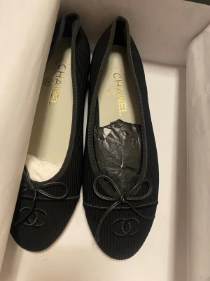 Svarta ballerinaskor från Chanel äkta skinn - Snygga svarta ballerinaskor från Chanel med klassisk rund tå och dekorativ rosett framtill. Skorna har diskret Chanel-logga broderad på tån och är tillverkade i läder med en elegant finish. Perfekta för dig som gillar tidlös och lyxig stil.