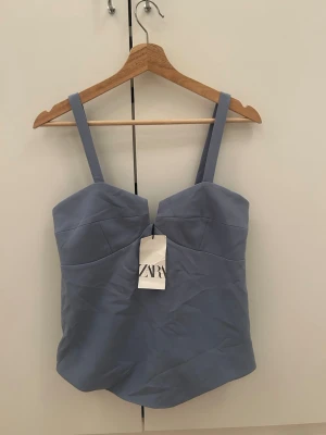 Blå bustier topp från Zara - Trendig blå bustier topp från Zara med breda axelband och en snygg v-ringad detalj framtill. Toppen har en figurnära passform och är tillverkad i ett slätt, mjukt material som ger en stilren look. Perfekt att matcha med jeans eller kjol.