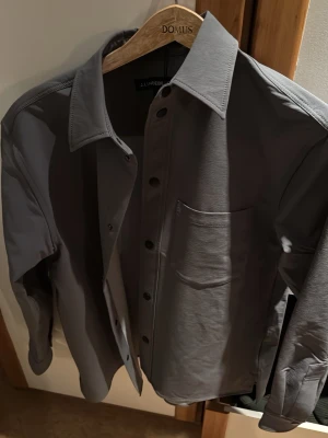 Grå skjorta från J.Lindeberg - Stilren grå skjorta från J.Lindeberg med klassisk krage, bröstficka och knappar framtill. Skjortan har lång ärm och en clean look som passar till många olika stilar. Perfekt för dig som gillar minimalistiskt mode. Helt ny och oanvänd. S men sitter som M.