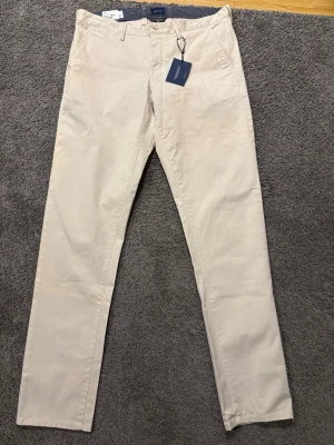 Beige slim GANT chinos 34/34 - Snygga beigea Gant chinos i slim fit-modell med twillstruktur. Klassisk design med fickor fram och bak, perfekt för en stilren look. Byxorna är aldrig använda och har kvar prislappen sedan köp. Storlek 34/34. Skriv gärna vid frågor och funderingar!😃✅