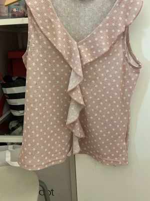 Rosa prickig volangtop från XOXO Friends - Söt ärmlös topp i ljusrosa med vita prickar från XOXO Friends. Toppen har en bred volangkrage och dekorativ volang framtill. Tillverkad i mjukt, ribbat material som känns bekvämt mot huden. Perfekt för en trendig och lekfull stil.