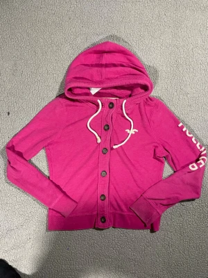Rosa hoodie från Hollister med knappar - Snygg rosa hoodie från Hollister med knappar framtill och vit dragsko i huvan. Hollister-logga på bröstet och tryck på ena ärmen. Tillverkad i mjukt bomullsmaterial, perfekt för en avslappnad och trendig look.
