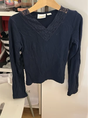 Mörkblå långärmad topp med spets - Mörkblå långärmad topp med snygg v-ringning dekorerad med spetsdetaljer. Toppen har en smal passform och är perfekt för dig som gillar stilrena plagg med en twist. Tillverkad i mjukt material som känns skönt mot huden.