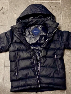 Mörkblå Gant jacka - Snygg mörk blå täckjacka från Gant med huva och två fickor (dragkedja) framtill, inklusive en mindre ficka innuti på vänstra sidan. Riktigt bra kvalitet på jackan knappast använd, inga skador. Håller värme bra under vintern men går fortfarande att använda under höst och vår.