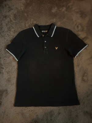 Svart piké från Lyle & Scott - Klassisk svart pikétröja från Lyle & Scott med korta ärmar och vit kant på krage och ärmslut. Tröjan har knappslå framtill och den ikoniska gula loggan på bröstet. Perfekt till en clean och stilren look.