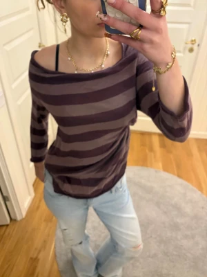 Randig offshoulder topp i lila toner - Säljer en randig långärmad topp i mörklila och ljuslila med offshoulder-design. Toppen har en avslappnad passform och är perfekt för en chill och trendig look. Materialet känns mjukt och skönt mot huden.