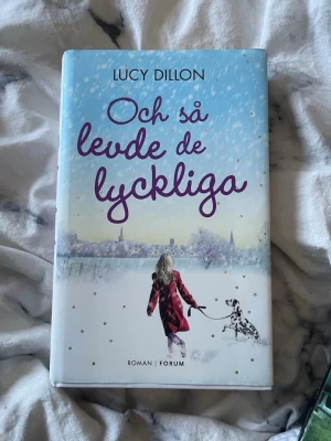 Och så levde de lyckliga av Lucy Dillon - Romanen 'Och så levde de lyckliga' av Lucy Dillon är en inbunden bok med ett omslag i ljusa blå och vita toner. Omslaget visar en kvinna i röd kappa som promenerar med en dalmatiner i ett vintrigt landskap. Perfekt för dig som gillar hjärtevärmande berättelser.