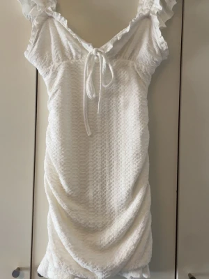 Vit kortklänning med volang och knyt - Supersöt vit kortklänning med volangdetaljer vid axlarna och knyt framtill. Klänningen har en strukturerad, vågig yta och är lätt rynkad i nederkant. Perfekt för sommarens alla tillfällen och ger en romantisk vibe.