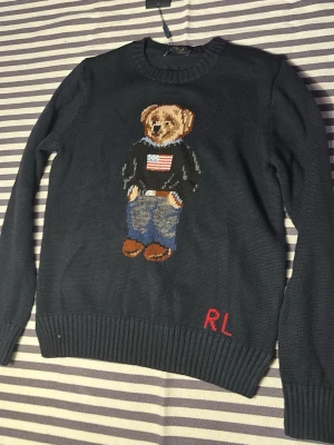 Mörkblå Polo Ralph Lauren stickad tröja - Mörkblå stickad tröja från Polo Ralph Lauren med ikonisk björn på bröstet, klädd i jeans, boots och tröja med amerikansk flagga. RL-broderi i rött nertill. Rund hals och ribbade muddar. Perfekt för dig som gillar klassisk preppy-stil.