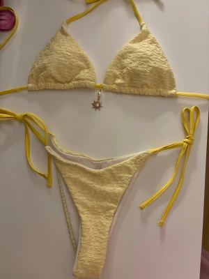 Gul bikini - Söt gul bikini med knyt i sidorna samt i nacken. Överdelen har trekantsform och ett solformat smycke framtill med tunna band för justerbar passform. Perfekt för strandhäng och soliga dagar! Den är oanvänd.