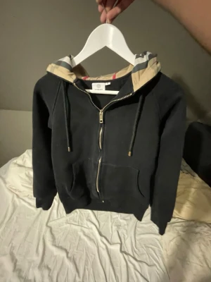 Svart zip hoodie från Burberry - Snygg svart hoodie från Burberry med dragkedja framtill och klassisk beige-rutig huva. Hoodien har justerbara snören i huvan och en stor ficka på magen. Perfekt för dig som gillar exklusiva streetwear-plagg och vill ha något stilrent med lyxig känsla.