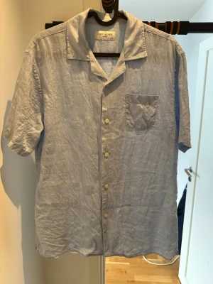 Ljusblå linneskjorta från John Henric - Säljer en ljusblå skjorta från John Henric i svalt linne. Skjortan har korta ärmar, klassisk krage, bröstficka och knappar framtill. Perfekt för varma dagar och enkel att styla till olika outfits. Endast använd ett fåtal gånger, 100% linne 